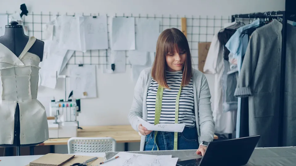 Empreendedora planejando o negócio em um ateliê de roupas, com croquis, fita métrica e notebook