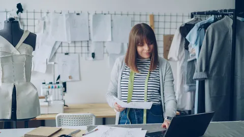 Empreendedora planejando o negócio em um ateliê de roupas, com croquis, fita métrica e notebook